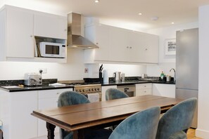 A modern kitchen with white cabinets, a stainless steel range hood, a microwave, a stove, and a dining area with a wooden table and chairs. - Charming 1BDR Retreat with Garden in West London (London)
