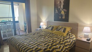 Ein Hotelzimmer mit Bett, Nachttisch, Lampe, einem Bild an der Wand und einem Balkon mit Stühlen.