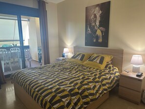 A hotel room with a bed, a nightstand, a lamp, a painting on the wall, and a balcony with chairs. - Beautiful apartment 85m2, with terrace in a +++ site, 5 swimming pools; 1 k from the sea (Manilva)