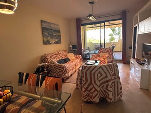A living room with patterned sofas, a glass coffee table, a dining table with a basket of fruit, and a painting on the wall. - Beautiful apartment 85m2, with terrace in a +++ site, 5 swimming pools; 1 k from the sea (Manilva)