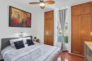 Ein Schlafzimmer mit einem Bett, Deckenventilator, einem Gemälde, Holzschränken und einem Fenster mit Vorhängen.