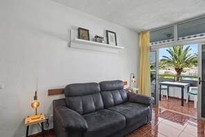 Ein Wohnzimmer mit Sofa, einem kleinen Tisch, einer Lampe, einem Regal mit Bildern und Blick ins Freie.