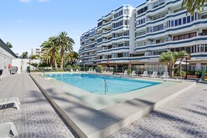 Una piscina con lettini, un percorso pedonale piastrellato e un edificio a più piani con balconi.