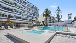 Ett poolområde på taket med en rektangulär pool, solstolar och utsikt över en hög byggnad med balkonger och en skylt.