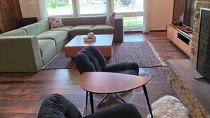 Uma sala de estar moderna com um sofá seccional, uma mesa de centro, uma poltrona preta, uma lareira e janelas amplas.