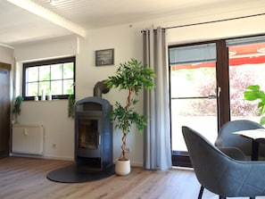 A room with a wood-burning stove, a dining table with chairs, a potted plant, and a window with curtains. - Quiet vacation apartment on the edge of the forest with panoramic views directly on hiking trails (30 m) (Prümzurlay)