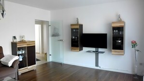 Una sala de estar moderna con un televisor de pantalla plana en la pared, armarios de madera con puertas de vidrio, un sofá y una pequeña mesa con un jarrón.