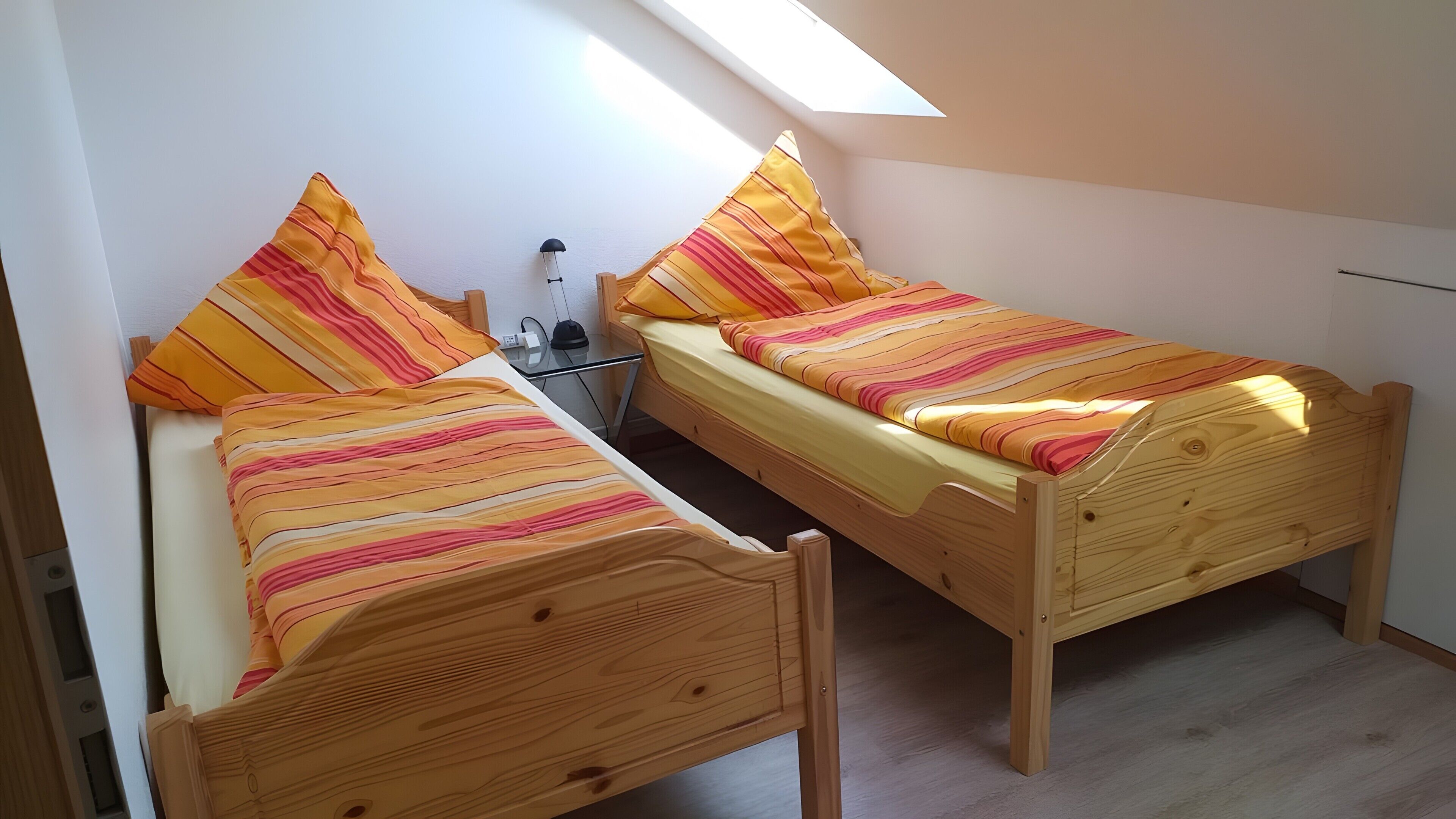 Ein kleines Zimmer mit zwei Holzbettgestellen, jeweils mit gestreifter Bettwäsche, einem kleinen Tisch mit einer Lampe und einem Oberlicht.
