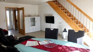 Ein Zimmer mit einer Holztreppe, einem an einer Backsteinwand befestigten Fernseher, einer Glasschrank und einem Tisch mit Dokumenten und einem Metallgestell.
