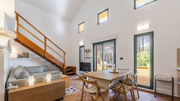 A modern living space with a wooden staircase, a sofa, a dining table with chairs, and a view through large windows. - Villa Dita Qerret | Relaxing Beachfront Stay by PikHost (Qerret)
