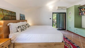 A bedroom with a bed, a nightstand, a wall-mounted lamp, a painting, an air conditioning unit, a sliding glass door, and a green cabinet with books. - Villa Dita Qerret | Relaxing Beachfront Stay by PikHost (Qerret)