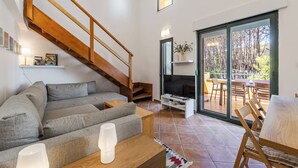 A living room with a grey sofa, wooden staircase, TV stand, and a sliding glass door leading to an outdoor patio. - Villa Dita Qerret | Relaxing Beachfront Stay by PikHost (Qerret)