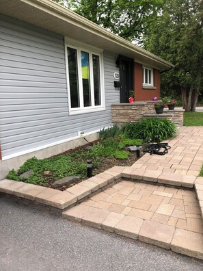 Una casa residencial con revestimiento gris, sección de ladrillo, un porche con plantas en macetas y un jardín cuidado.