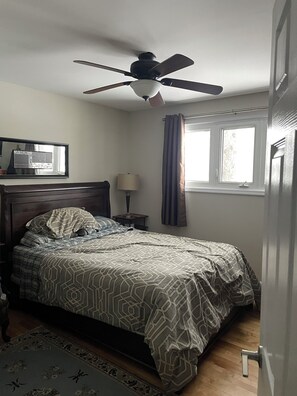 Un dormitorio con cama, ventilador de techo, ventana con cortinas, mesita de noche con lámpara y espejo en la pared.