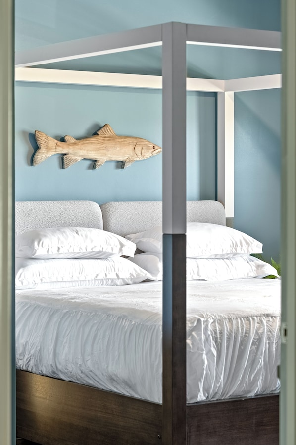 A four-poster bed with white bedding, a wooden fish wall art, and a blue wall.