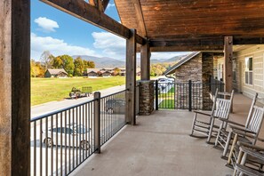 A covered porch with a view of a grassy field and houses, wooden chairs, and a stone pillar.