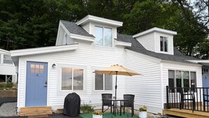 A white single-story house with a blue door, a patio with a table and chairs, and a covered area with an umbrella. - Incredible Lake & Mtn Views - Loft Vibes Getaway! (Tilton)