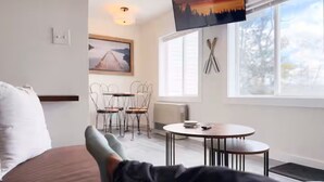 A bed with a pillow, a person's legs, a table with chairs, a painting, and a window with a view. - Incredible Lake & Mtn Views - Loft Vibes Getaway! (Tilton)