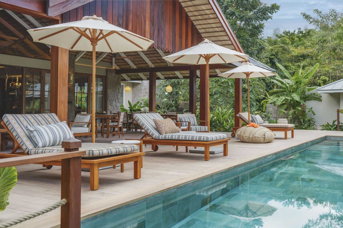 Zona junto a la piscina con sillones de descanso y sombrillas, rodeada de exuberante vegetación.