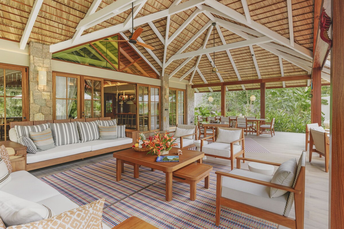 Amplio espacio interior con techo alto, vigas de madera y techo de paja. Cuenta con una zona de estar con sofá y sillones, una mesa de centro con adornos, y vistas a exuberante follaje exterior.