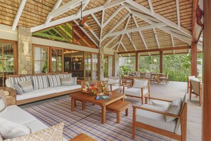 Amplio espacio interior con techo alto, vigas de madera y techo de paja. Cuenta con una zona de estar con sofá y sillones, una mesa de centro con adornos, y vistas a exuberante follaje exterior.