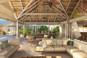 An outdoor lounge area with a high ceiling, wooden beams, and a thatched roof. There are multiple seating areas, including a sofa and chairs, and a dining table with chairs.