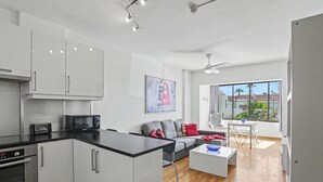 Een moderne keuken met witte kastjes, een zwart aanrechtblad en een magnetron. Er is een grijze bank met rode kussens, een witte salontafel en een eethoek met een tafel en stoelen. Een groot raam biedt uitzicht op buiten.
