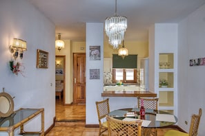 A dining area with a chandelier, wooden chairs, a glass table, and a kitchen in the background.