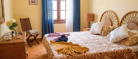 Habitación de hotel con una cama, una silla, una mesa con un jarrón de flores, una lámpara y un ventanal con cortinas.