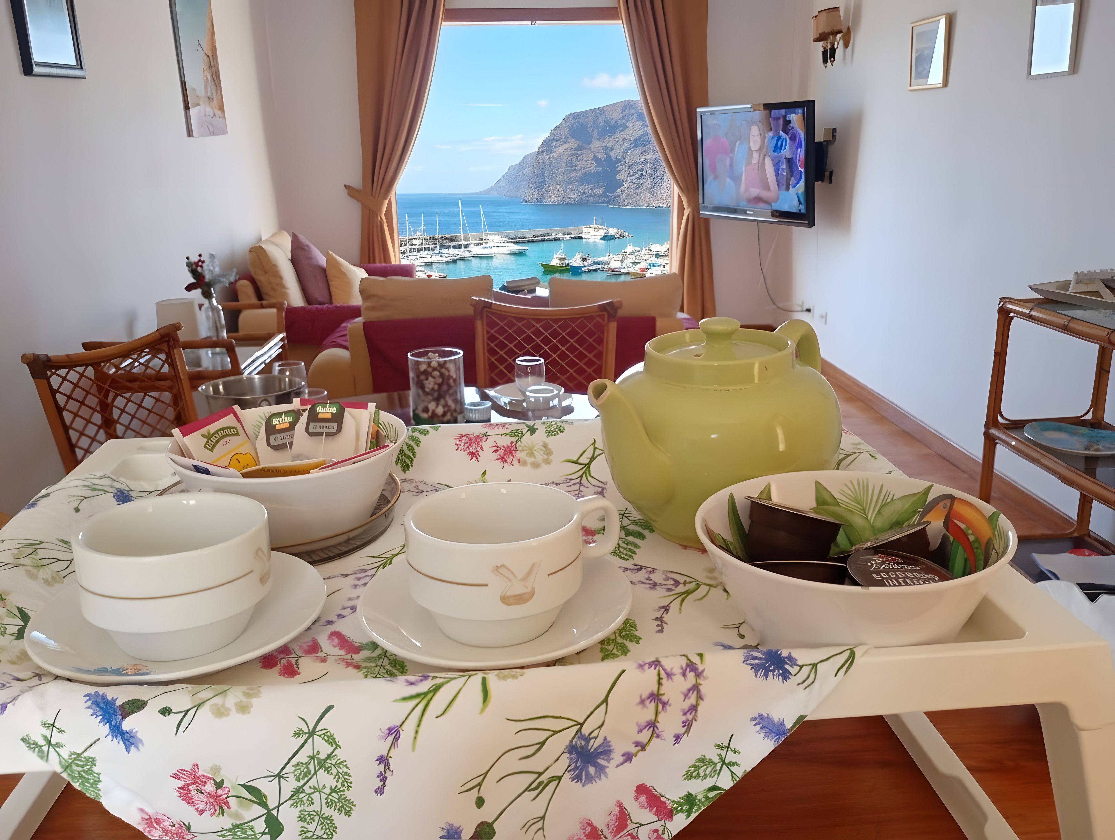 A dining area with a table set for tea, a view of the sea, and a television on the wall.
