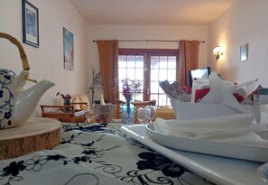 A hotel room with a dining table set, a vase with flowers, a wooden side table, and a window with curtains.
