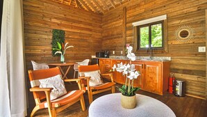 Interno di una baita in legno con un tavolo rotondo, due sedie, un angolo cottura e una finestra che si affaccia sul verde.