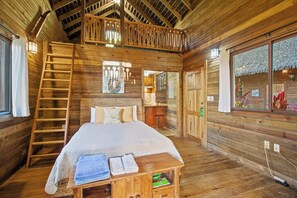 A wooden cabin with a loft bedroom, a ladder to the upper room, a bed with pillows, a small table with towels, and a window with a view.