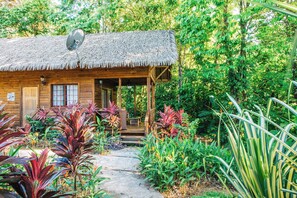 A wooden cabin with a thatched roof, surrounded by lush greenery and a variety of plants.