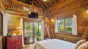 Ein Schlafzimmer in einer Holzhütte mit einem Bett, einem Schreibtisch mit Lampe, einem Fernseher und einem Balkon mit Blick auf Grünflächen.