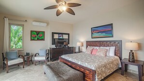 Een slaapkamer met een groot bed, een plafondventilator, een dressoir, een bureau, twee stoelen, een bankje en een raam met uitzicht op groen.
