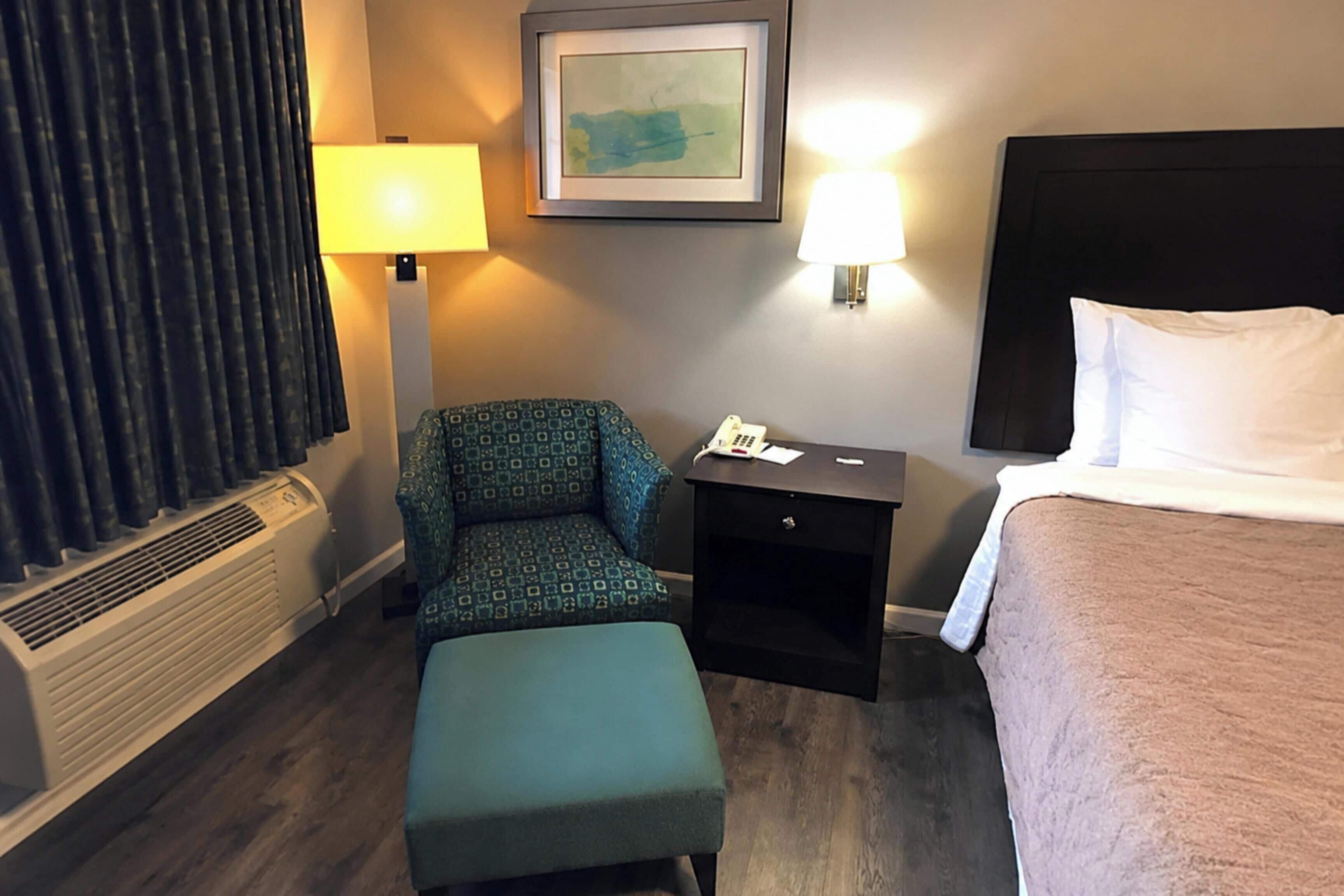 A hotel room with a bed, a chair, a bedside table with a lamp, a heater, and a framed picture on the wall.