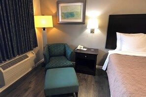 Habitación de hotel con una cama, una silla, una mesita de noche con lámpara, un calentador y un cuadro en la pared.