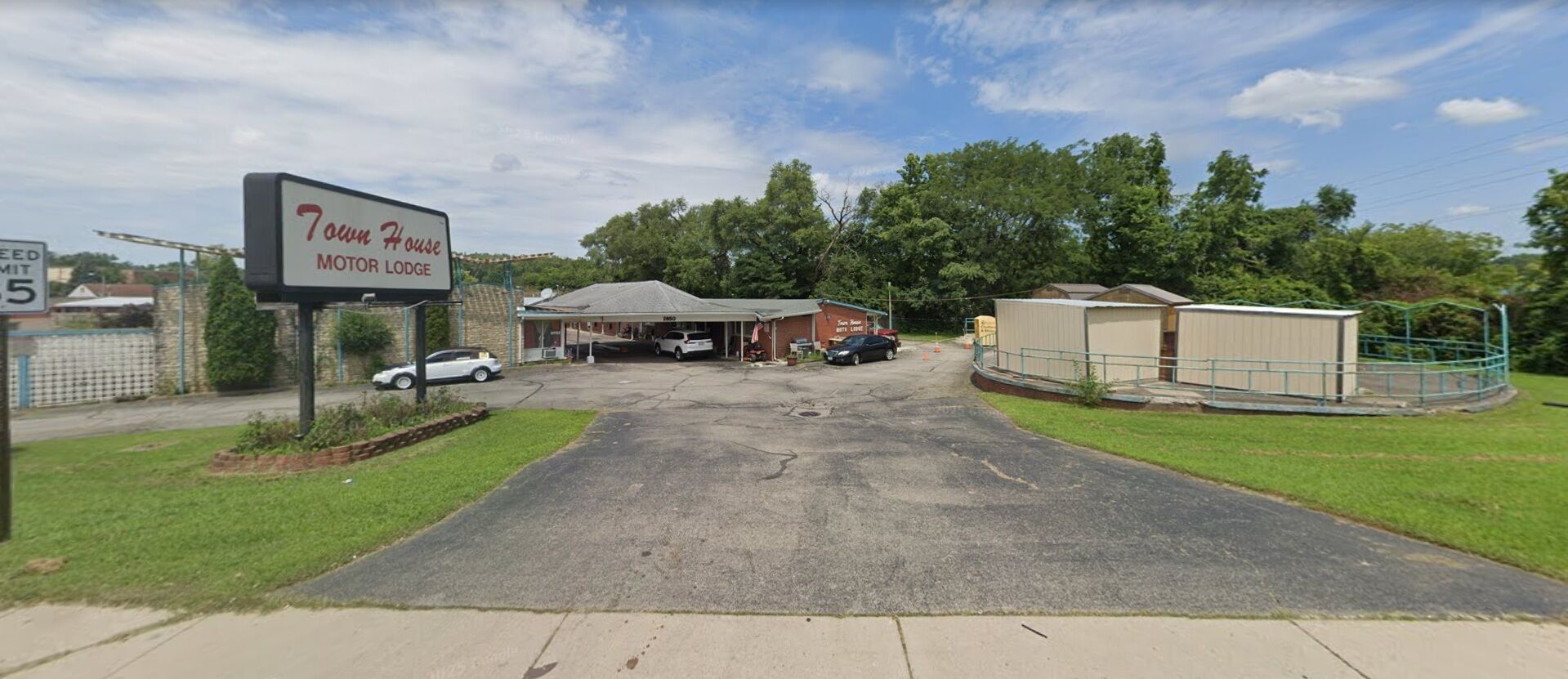 A motel with a sign reading '7own House Motor Lodge,' a parking lot, and a fenced area with buildings.