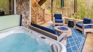 A hot tub with a stone wall and wooden deck, surrounded by blue cushioned chairs and a fire pit. - Luxury Mountain Estate | Spectacular Views (Sweden)