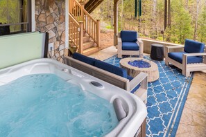 A hot tub with a stone wall and wooden deck, surrounded by blue cushioned chairs and a fire pit. - Luxury Mountain Estate | Spectacular Views (Sweden)