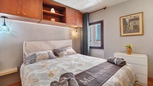 Letto rifatto con copriletto a tema 'Vista Museo', pensili in legno sopra, un tavolino con un vaso e un quadro appeso al muro.