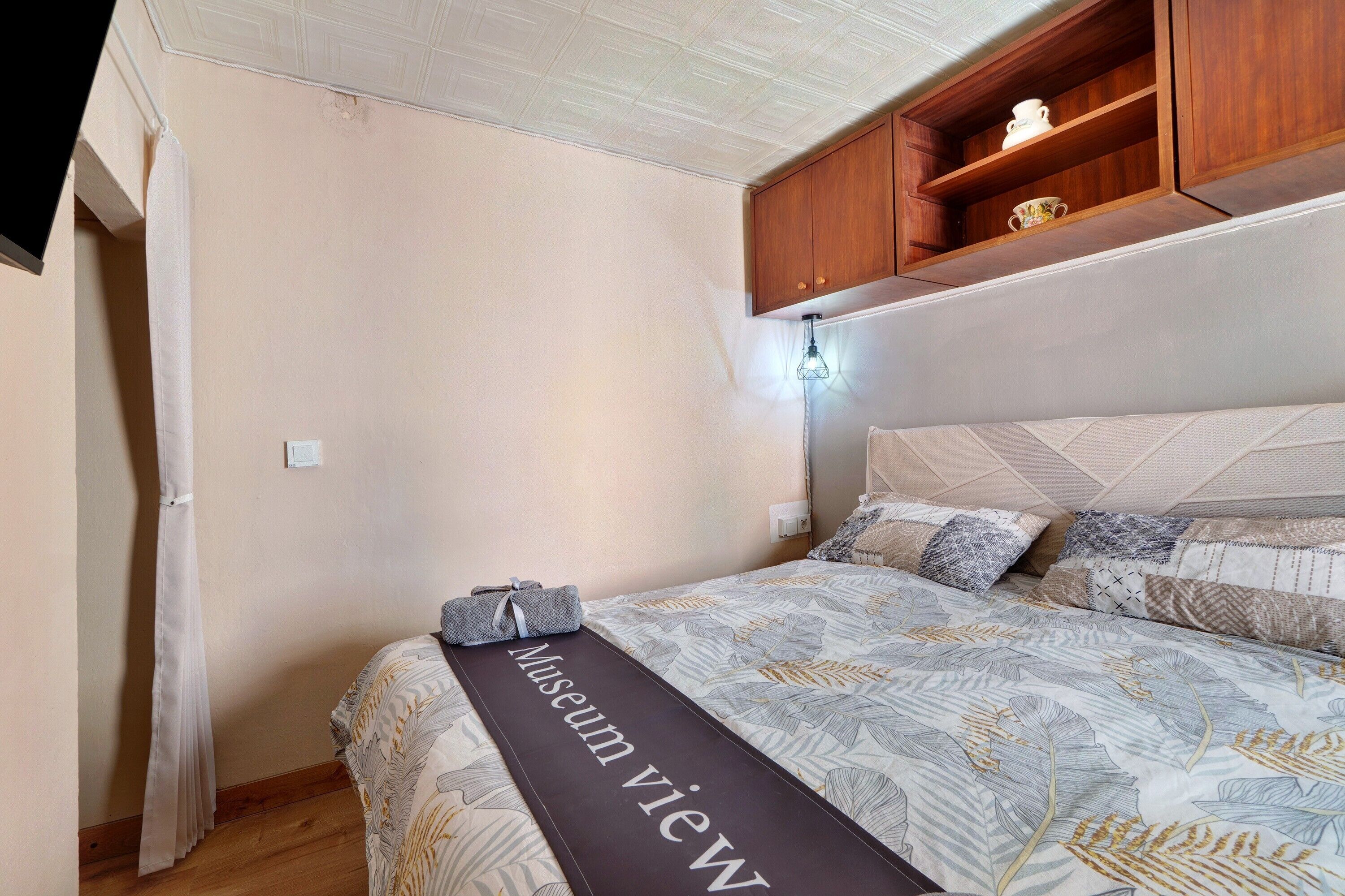 Un dormitorio con cama, estante de madera sobre la cama, mesita de noche con lámpara y una toalla en el suelo.