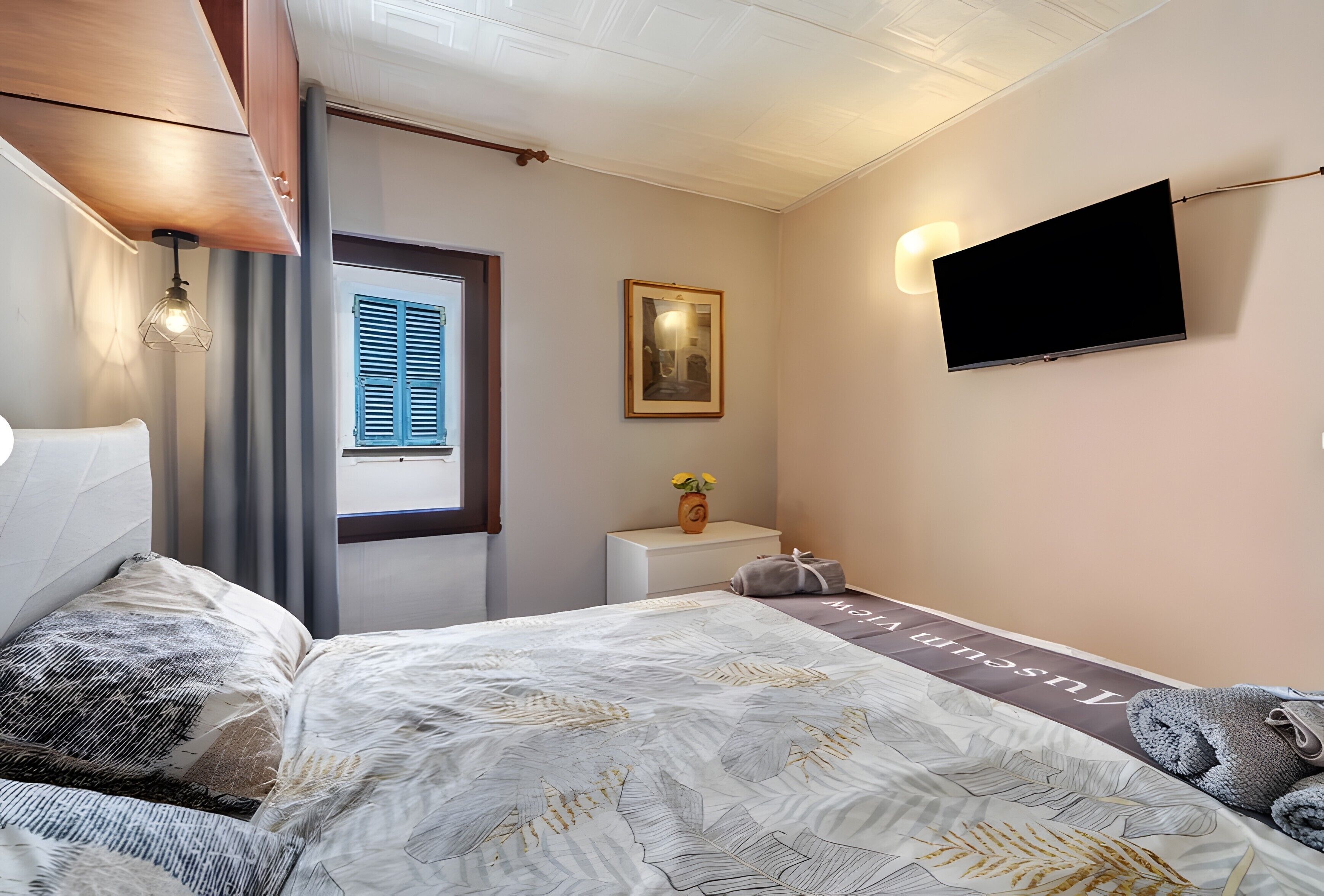 Ein Schlafzimmer mit Bett, Nachttisch, Fernseher, einem gerahmten Bild und einem Fenster mit Rollläden.