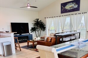 Ein Wohnzimmer mit Fernseher, Esstisch, Sofa, Deckenventilator und einem großen Fenster mit Vorhängen.