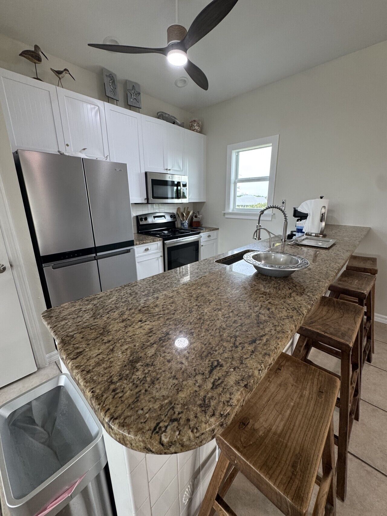 A kitchen with granite countertops, stainless steel appliances, wooden bar stools, and a ceiling fan.