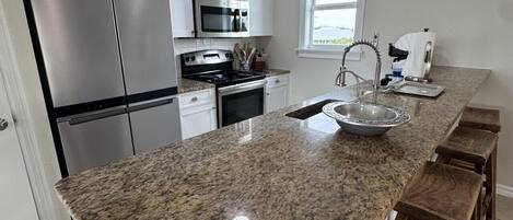 A kitchen with granite countertops, stainless steel appliances, wooden bar stools, and a ceiling fan.