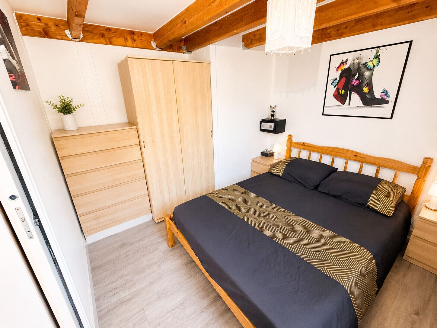 Een slaapkamer met een bed, een houten kledingkast, nachtkastje, lamp, radio en een ingelijst schilderij aan de muur.