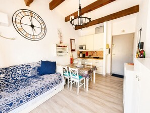 Ein Zimmer mit einer gemusterten Couch, einem Esstisch mit Stühlen, einer Küchenzeile und einer großen Wanduhr.
