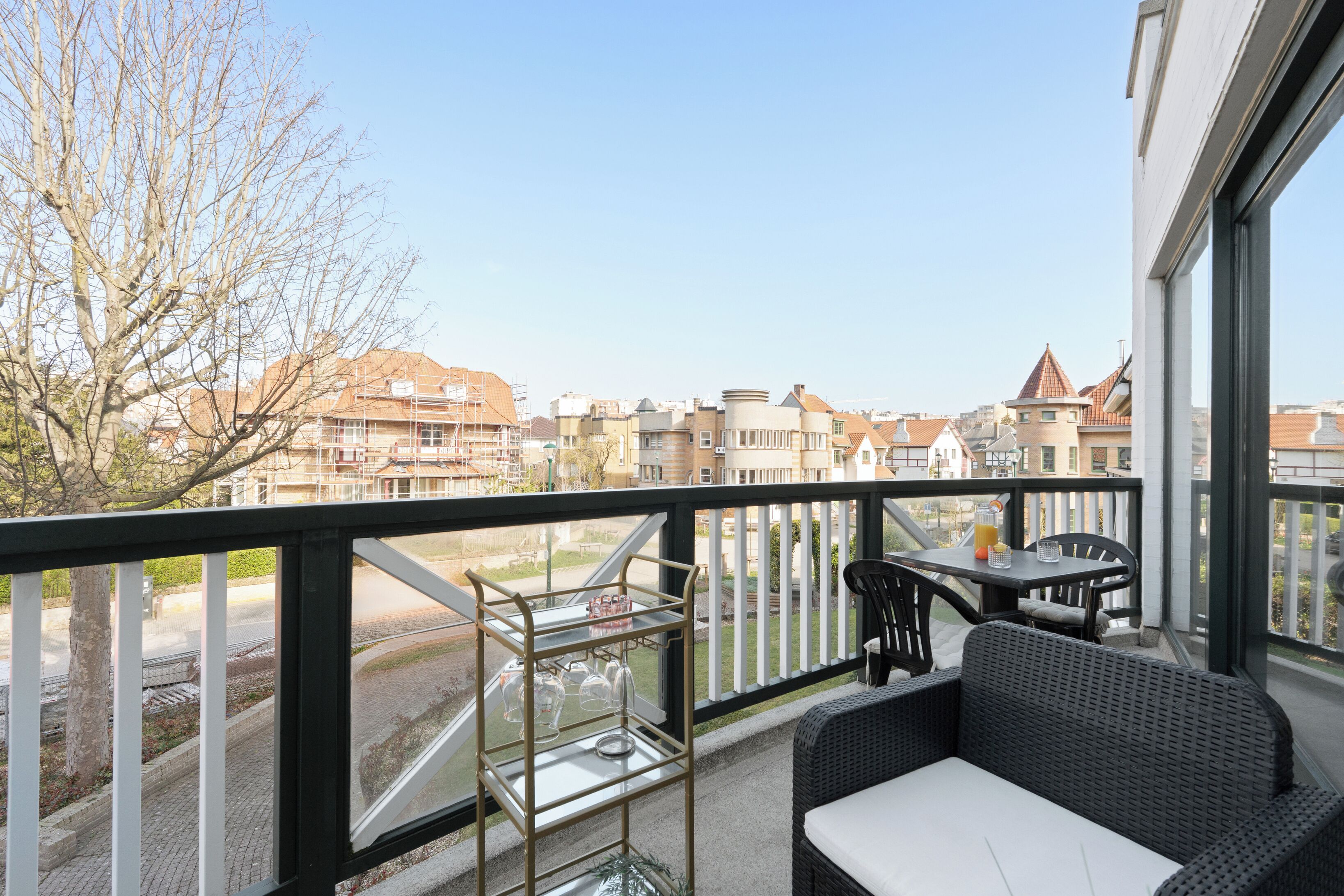 A balcony with a view of buildings, a glass table with chairs, and a metal wine rack.